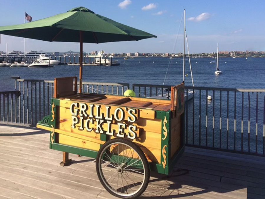 A wooden cart with an umbrella labeled "Grillo's Pickles" by a waterfront.