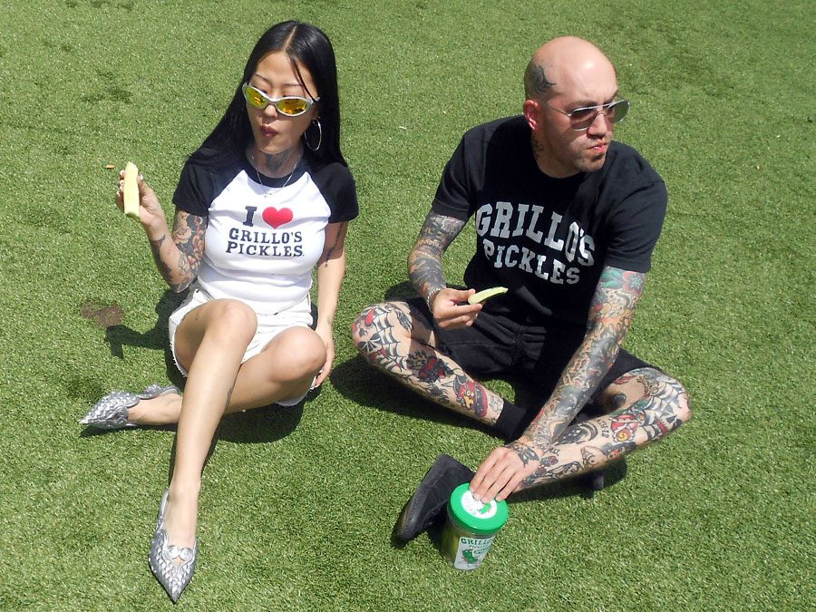 A man and a woman sit on green grass, enjoying pickles. They wear  Grillo's Pickles shirts and sunglasses.